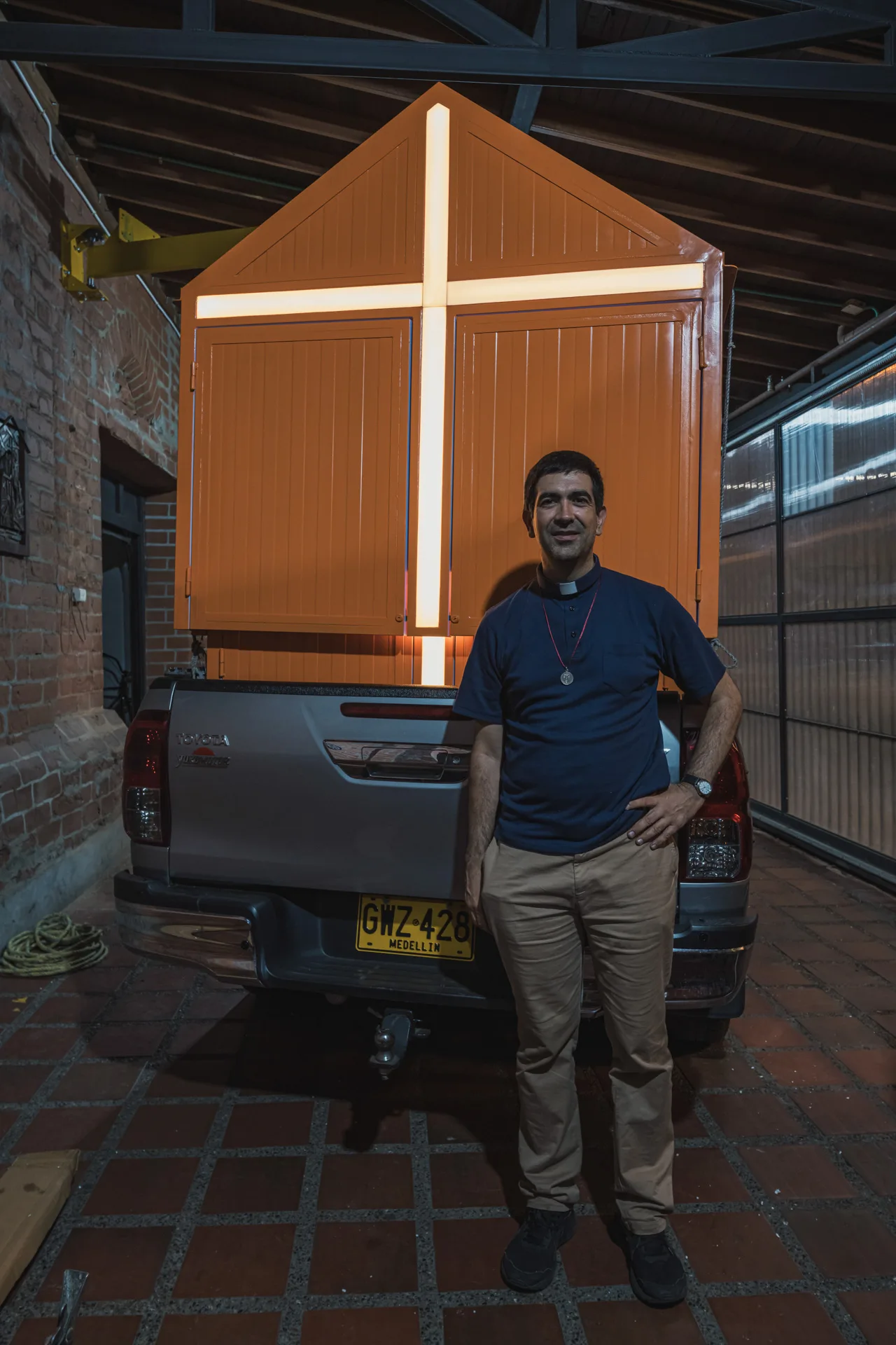 Confesionario móvil de A5 Arquitectura con una cruz luminosa, montado sobre un camión pick-up en Medellín, junto a un sacerdote.